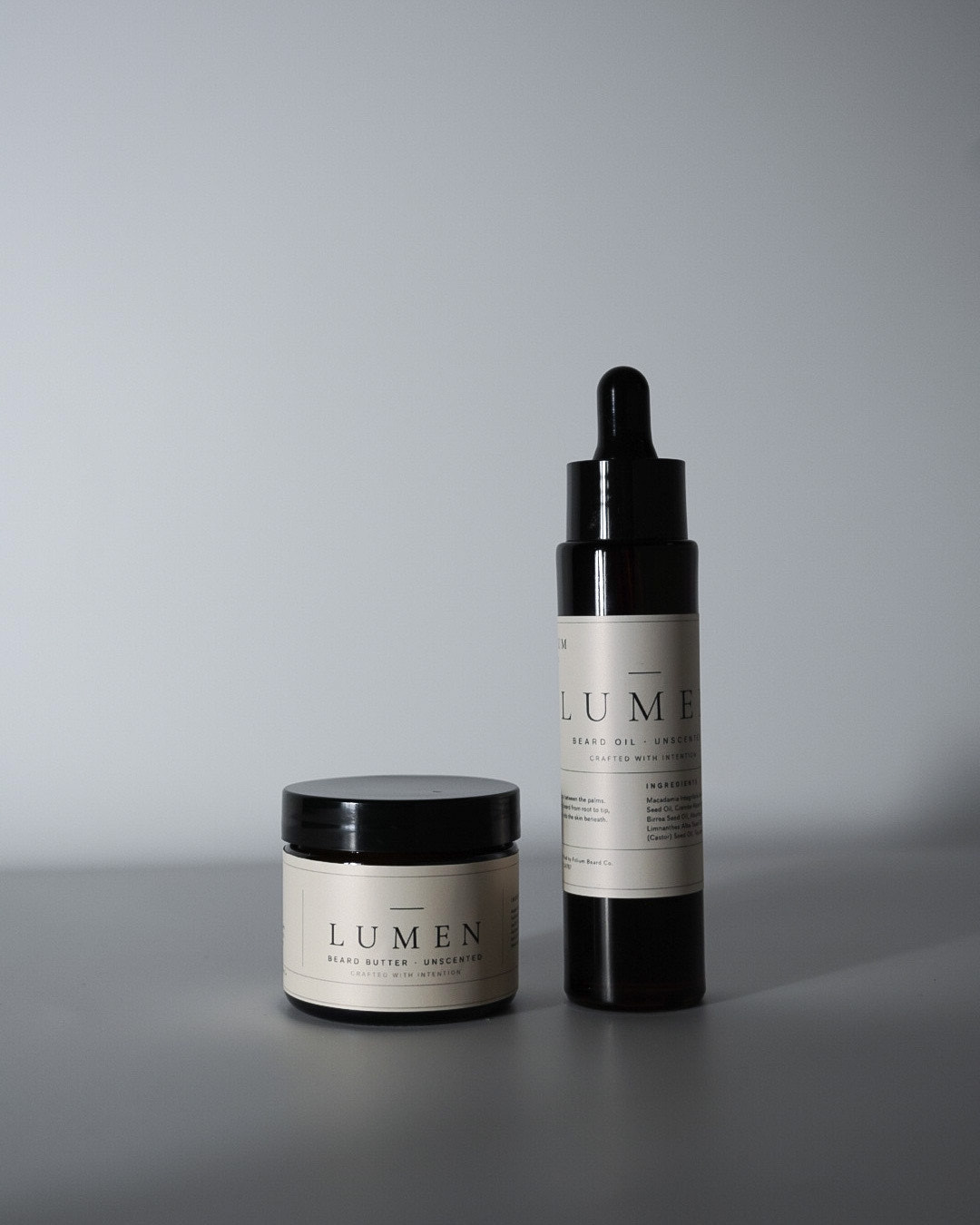 LUMEN unscented beard oil and beard butter set in amber glass packaging on a soft gray background with natural directional light.