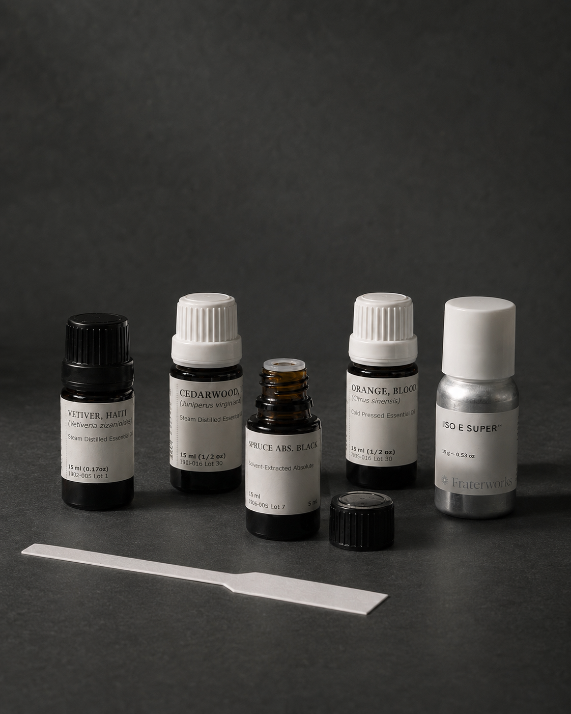 Folium Beard Co. fragrance materials on a dark grey surface — steam-distilled vetiver and cedarwood essential oils, a solvent-extracted spruce absolute, cold-pressed blood orange, and synthetic Iso E Super, with a scent blotter.