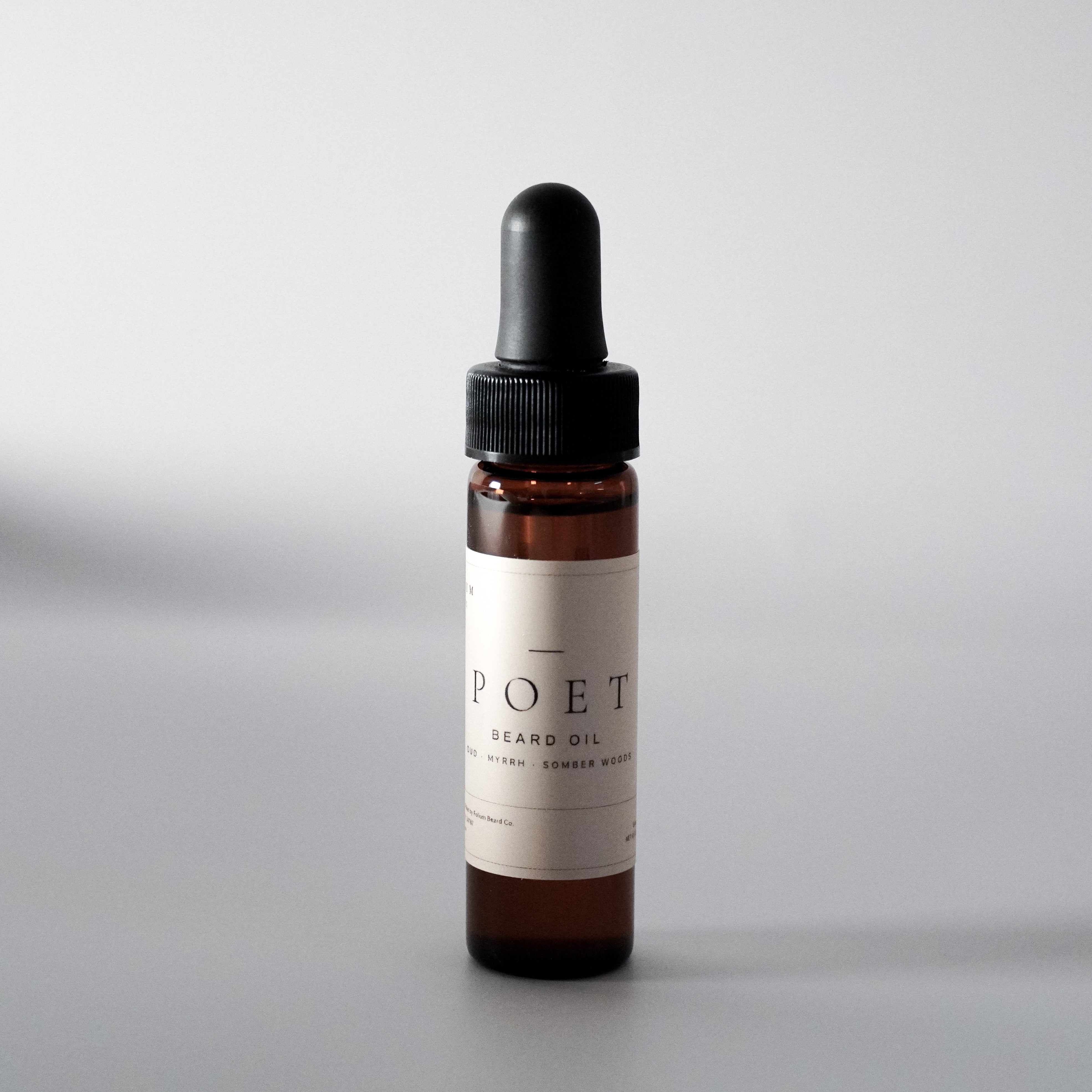 The Poet oud and myrrh beard oil in a 15 ml bottle on a cool grey background.